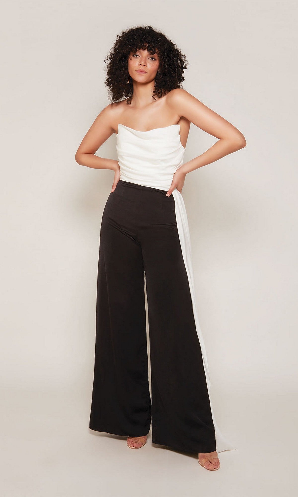 A woman stands with hands on hips, modeling the Alyce 70050 Long Formal Jumpsuit, which has a strapless white top and high-waisted black wide-leg pants with a white side panel, set against a plain light background.